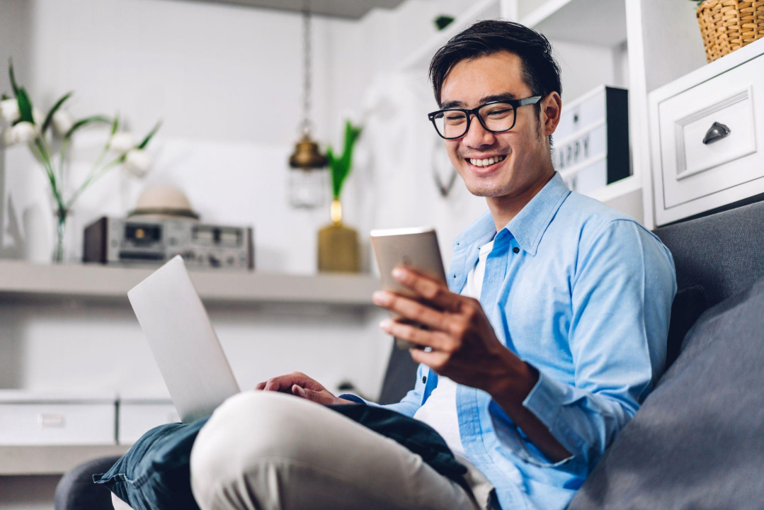 pria-duduk-di-sofa-dengan-laptop-bermain-handphone